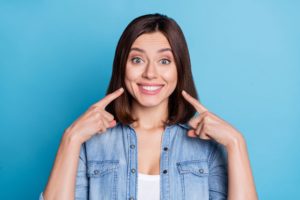 Woman with brown hair and a denim jacket in front of a blue background pointing to her white smile with 2 fingers